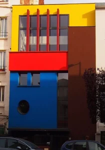 Maison de ville contemporaine aux façades colorées bleu, rouge, jaune et marron, avec hublot et grandes baies vitrées, conçue par Le Quilleuc Architecte en cœur d’îlot urbain.